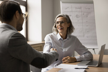 Cheerful mature business professional woman shaking hands with younger coworker man at laptop, smiling, laughing. Project partners giving greeting handshake after successful meeting