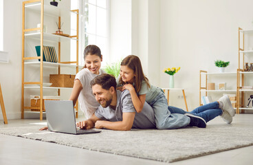 Happy young parents with their child girl lying on the floor at home using laptop. Smiling family...