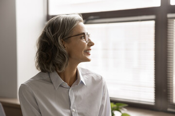 Happy thoughtful business leader woman in glasses looking at office window with toothy smile,...