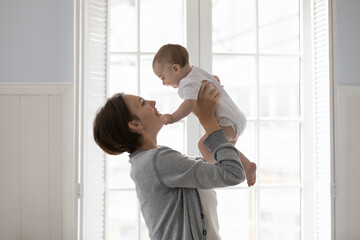 Fototapeta premium Happy young mother play with newborn at home, standing in cozy light domestic room, lifts, raise up on arms her adorable baby in bodysuit enjoy playtime, motherhood, time together. Babyhood, new life