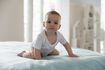 Fototapeta premium Beautiful newborn babygirl or babyboy in bodysuit awakened in cozy bedroom kneeling on white bedsheets, staring at camera. Carefree babyhood, healthcare, paediatrics, childcare, new Alpha generation