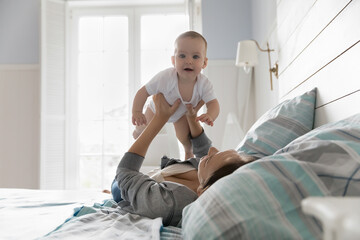 Loving young mother lifts her adorable newborn baby, express caress and tenderness, enjoy playtime and sweet moments, lying on comfortable bed in bedroom. Happy motherhood, cherish, unconditional love