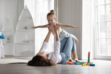 Happy young mother lying on warm floor in playroom, lifts little daughter, girl imitate airplane, imagining flying in air, carefree family enjoy active domestic games. Motherhood, relationships, bond - obrazy, fototapety, plakaty