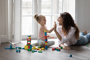 Happy mother playing wooden blocks with daughter, finish build tower, praising kid, giving high...