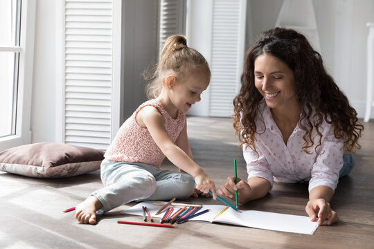 Caring babysitter drawing pictures with pencils spend time at home with little cute girl, settled down together on warm floor, enjoy creative hobby and communication. Family pastime, kid development