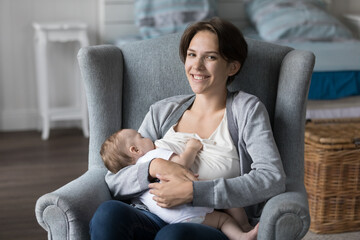 Portrait of loving mother sit on cozy armchair breastfeeding newborn in modern bedroom smile look...
