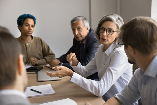 Confident mature business leader woman speaking to multiethnic diverse team of employees, giving instructions, explaining project plan, work strategy, offering ideas for brainstorming
