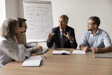 Serious senior business professional man in formal suit working with team, sitting at meeting table...
