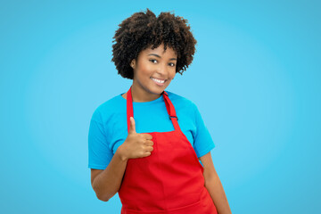 Happy african american waitress with red apron showing thumb up on blue background