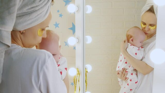 Young mother take care of her skin with under eye patches holding her newborn baby in her arms in front of the mirror at home. Motherhood concept.