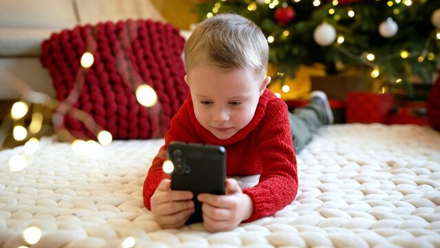 A boy lies on the floor at home and plays a computer game. New Year and Christmas, little boy on the floor. A little boy lies on the floor and looks at the phone, against the backdrop of a decorated.