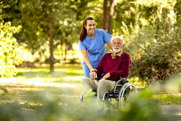 Female nursing assistant and elderly man in wheelchair enjoying autumn in the park and looking to the camera.