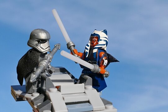 LEGO Star Wars Togruta female Jedi Ahsoka Tano fighting with Third Order stormtrooper captain Phasma, lightsabers against baton, standing on top of unfinished AT-ST battle droid. Blue skies. 