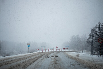 Photo of detour road signs covered with snow during the snowfall in Magadan, Russia. Snow showers on the trees and hills. Slush on the way. Fog and haze, low visibility due to snowstorm. Extreme