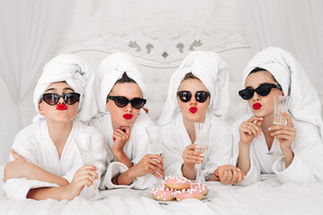 Young, beautiful women in bathrobes and glasses enjoying bachelorette party at spa or hotel and eating donuts, happy multiracial girlfriends celebrating bachelorette party