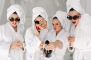 Photo with champagne opening and a flying cork. Women in bathrobes and glasses enjoy their...