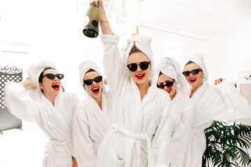Cheerful cute multiracial young girlfriends in bathrobes having fun, holding glasses of champagne, sitting on bed in hotel. Concept for women's relaxation, spa and entertainment. - obrazy, fototapety, plakaty