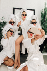 Cheerful cute multiracial young girlfriends in bathrobes having fun, holding glasses of champagne,...