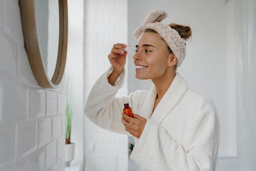 Beautiful young woman in bathrobe applying cosmetic serum on face while standing in bathroom