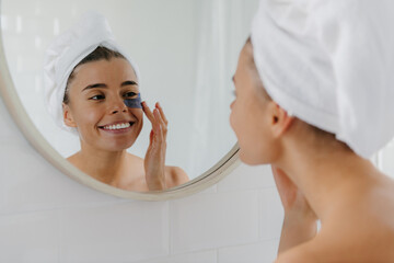 Smiling young woman covered in towel applying eye patches while looking at the mirror in bathroom