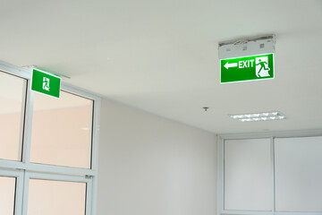 Selective fire exit sign on white ceiling and glass door.Green fire escape sign hang on the ceiling...