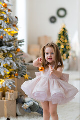 Smiling little girl in an elegant dress holds a Christmas tree toy near the Christmas tree, celebrating the New Year at home