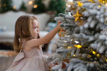 Pensive little girl in an elegant dress holds a Christmas tree toy near the Christmas tree,...