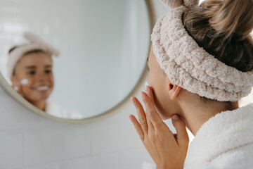 Beautiful young woman in bathrobe applying face cream while looking at her reflection in the mirror - 672722376
