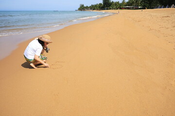 A woman sits with her back to the sea and is writing the letters "L" "O" to express the word LOVE on the fine sand. Female tourists drawing in the sand happily at the beach while visiting the sea.
