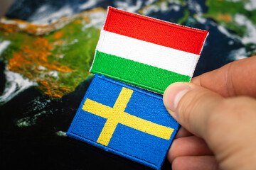 Fototapeta premium small flags of Sweden and Hungary held in hand against the background of a world map, Concept, mutual relations of both countries, Hungary blocking Sweden's accession to the defense pact