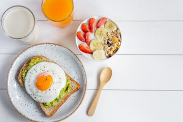 Healthy Breakfast food. Avocado toast with fried egg and bowl of yogurt with granola ,banana, strawberry. And Fresh orange juice and milk on white wooden, Top view ,Copy space.