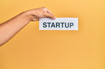 Hand of caucasian man holding paper with startup word over isolated yellow background