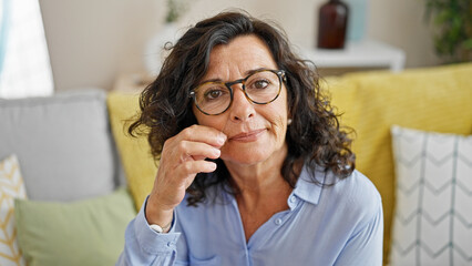 Middle age hispanic woman sitting on sofa doing silent gesture at home