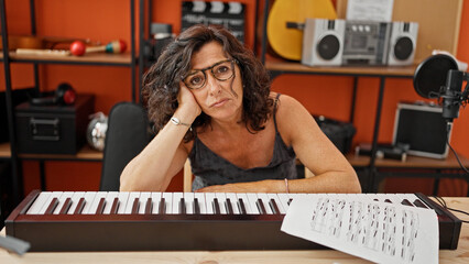 Middle age hispanic woman musician leaning on piano with sad expression at music studio