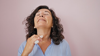 Middle age hispanic woman smiling confident massaging face with skin roller over isolated pink background