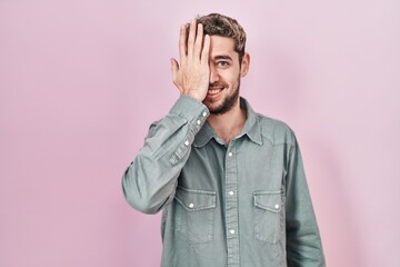 Hispanic man with beard standing over pink background covering one eye with hand, confident smile on face and surprise emotion.