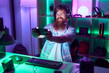 Young redhead man streamer playing video game using smartphone at gaming room