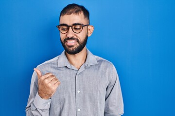 Middle east man with beard standing over blue background smiling with happy face looking and pointing to the side with thumb up.