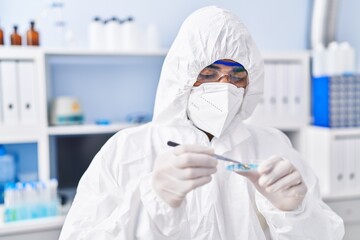 Young hispanic man scientist wearing medical mask holding sample with tweezers at laboratory