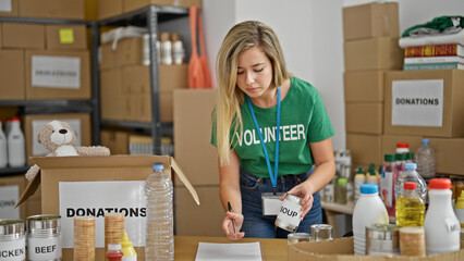 Young blonde woman volunteer writing on document checking products at charity center