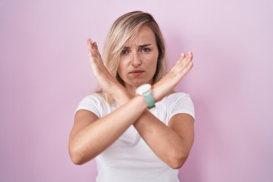 Young blonde woman standing over pink background rejection expression crossing arms doing negative sign, angry face