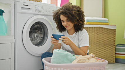 Young beautiful hispanic woman playing video game waiting for washing machine at laundry room