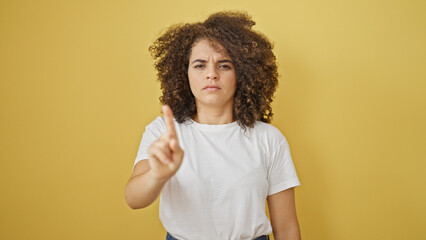 Young beautiful hispanic woman standing with serious expression saying no with finger over isolated yellow background - obrazy, fototapety, plakaty