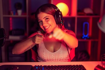 Young blonde woman playing video games wearing headphones pointing fingers to camera with happy and...