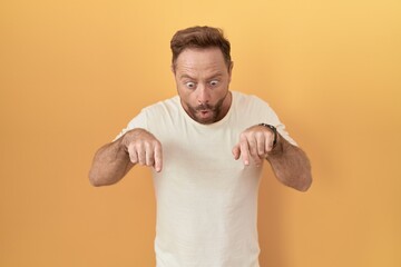 Middle age man with beard standing over yellow background pointing down with fingers showing advertisement, surprised face and open mouth