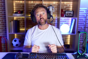 Middle age man with beard playing video games wearing headphones making fish face with lips, crazy...