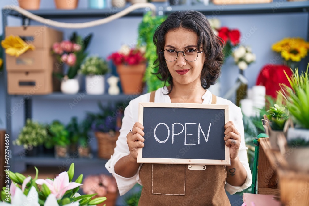 Sticker young hispanic woman working at florist holding open sign clueless and confused expression. doubt co