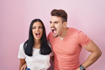 Young hispanic couple standing over pink background angry and mad screaming frustrated and furious, shouting with anger. rage and aggressive concept.