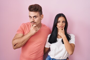 Young hispanic couple standing over pink background looking confident at the camera smiling with crossed arms and hand raised on chin. thinking positive.