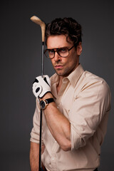 Portrait of stylish and serious man in shirt and eyeglasses holding golf club isolated on grey 